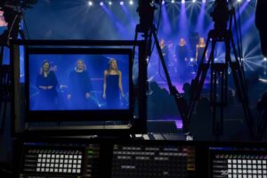 Ekran podglądowy ze stanowiska realizacyjnego, pokazujący trzy wokalistki na scenie. Wokół ekranu widoczne statywy kamer telewizyjnych.