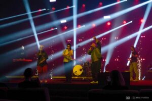 Zespół muzyczny (kilka osób) występuje na scenie, otoczonej mocnymi, białymi promieniami świetlnymi na czerwonym tle.