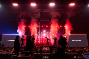 Scena z dwoma ekranami LED i dymem scenicznym, widziana z perspektywy stanowiska realizacyjnego z kamerzystami w pierwszym planie.