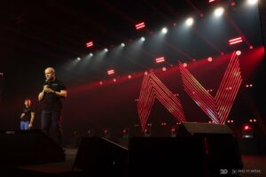 Piotr Łukasiewicz na scenie, w tle dynamiczna grafika AV i czerwone oświetlenie.