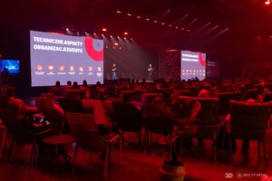 Prelegenci na scenie przed dużą publicznością, na wielkoformatowych ekranach LED wyświetlany jest slajd o technicznych aspektach organizacji eventu.