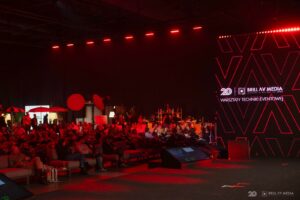 Widownia siedząca na wygodnych fotelach w dużej sali, na której dominuje scena z dużym ekranem LED z czerwonym logo Brill AV Media.